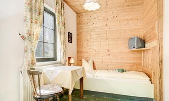 A cozy room with a wooden wall and a window. There is a table, a chair, and a single bed with simple bedding.
