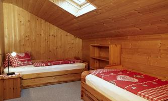 A cozy bedroom with two beds and wooden paneling. The room features a window with daylight and simple wooden furniture.