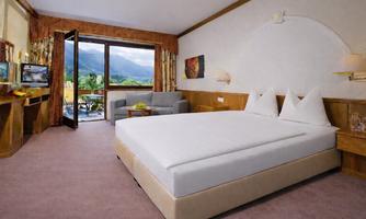A modern hotel room with a large double bed and cozy seating. The view from the window shows a picturesque mountain landscape.