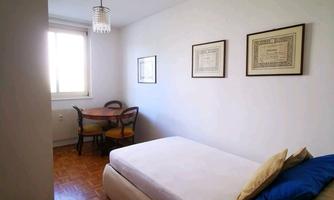 A simple bedroom with a single bed and a table. Two pictures are hanging on the wall and there is a window with plenty of natural light.