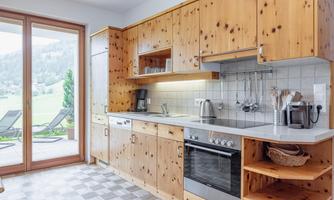 A modern kitchen with wooden cabinets and a large window. Through the window, a green landscape with loungers is visible.
