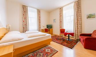 A bright and inviting room with two beds and a cozy armchair. The room is adorned with curtains and a carpet and offers a television and a small table.