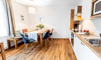 A modern kitchen with a dining table and chairs. The room is bright and friendly, with a wooden floor and flowers on the table.