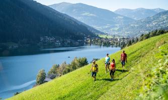 A group of hikers walks along a green meadow by the shore of a lake. In the background, mountains and a clear sky can be seen.
