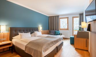 A modern hotel room with a double bed and bright wooden furniture. Large windows provide plenty of daylight and a cozy atmosphere.