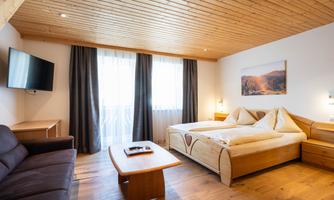A cozy hotel room with wooden furniture and a large window. There is a double bed, a couch, and a television.