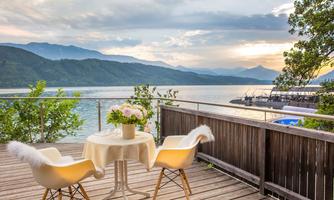 A cozy terrace with a table and two chairs, overlooking a peaceful lake and surrounding mountains. The sky is cloudy, and the sunset creates a relaxed atmosphere.