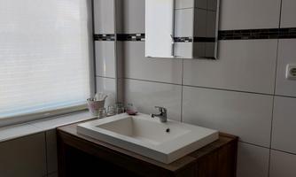 A modern bathroom with a sink on a wooden cabinet. There is a window with rollable blinds and bright tiled walls.