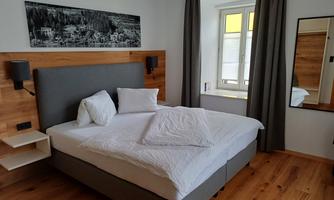 A modern bedroom with a large double bed and wooden floor. The walls are light, and there are windows with curtains as well as a mirror on the wall.