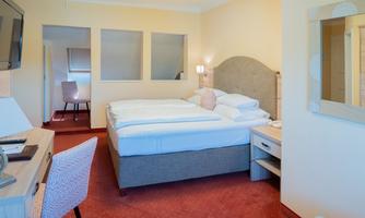 A bright, cozy hotel room with a double bed and a desk. The walls are painted in soft colors and there is a flat-screen TV.