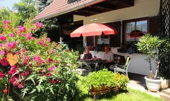A cozy garden in front of a house with colorful flowers and a sun-protecting umbrella. Two people are sitting relaxed at the table, enjoying their time outdoors.