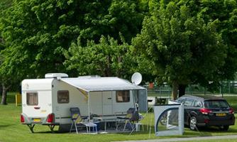 A caravan is standing on a spacious lawn area, surrounded by trees. A car is parked next to the caravan, and some garden chairs are set up.