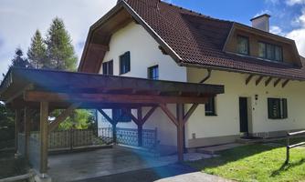 A modern house with a gabled roof and a covered terrace. The entrance area is surrounded by a green space.