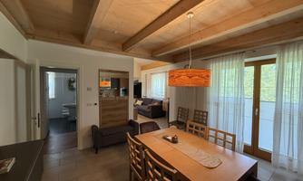 A cozy dining area with a large table and modern chairs. In the background, there is a bright living space with large windows and soft curtains.