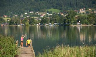 A picturesque landscape with a tranquil lake, surrounded by trees and hills. Two people stand on a pier and enjoy the beautiful nature.