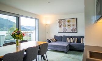 A modern living room with a large sofa and a dining table. Large windows offer a view of the surrounding landscape.
