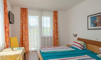 A bright bedroom with two beds and colorful bedding. The large windows let in a lot of daylight.
