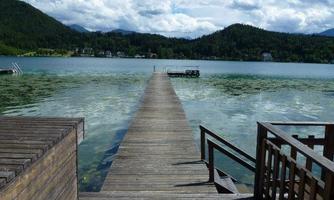 A wooden walkway leads over the clear water to a lake, surrounded by hills and a blue sky with white clouds. The tranquil landscape invites relaxation.