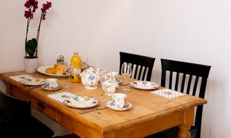 A beautifully set wooden table with tea cups, plates, and cutlery. There is also a fruit bowl and a carafe with drinks on it.