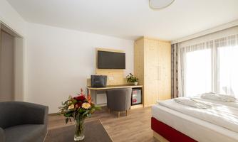 A modern hotel room with a bed, a table, a chair, and a television. Flowers in a vase adorn the table, and the room has large windows with naturally bright light.