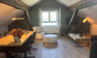A cozy attic room with wooden beams, a dining table, and modern furniture. A window lets in plenty of light and creates a friendly atmosphere.
