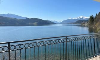 A peaceful lake view with mountains in the background and a clear sky. The view is from a balcony with a wrought-iron railing.