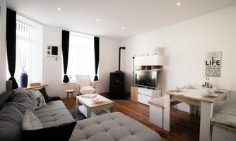 A modern living room with a gray sofa and a cozy dining area. There are large windows that let in plenty of light, and a stylish wall decoration.