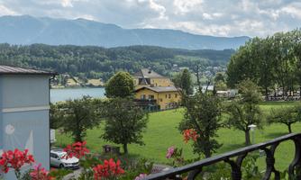 A picturesque view of a lake with a yellow house in the middle. In the foreground, colorful flowers are blooming, and in the background, mountains and forests can be seen.