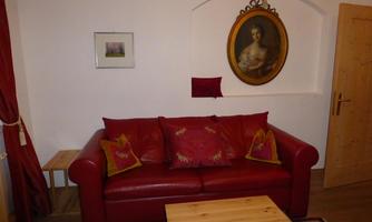 A cozy living room with a red sofa and colorful pillows. On the wall hangs an oval painting of a woman.