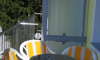 A small balcony with a round table and four yellow-orange chairs. In the background, green trees are visible.