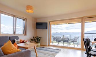 A modern living room with a cozy couch and a dining table. Large windows provide a beautiful view of the mountains and the balcony.