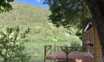 A wooden terrace by the shore of a clear lake, surrounded by green trees and mountains. The sky is blue and sunny.