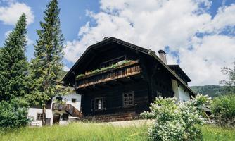 A traditional wooden house with a balcony and beautiful flower planting. Surrounded by green meadows and tall trees under a clear sky.