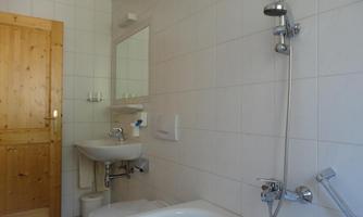 A simple bathroom with white tiles. Visible are a bathtub, a sink, and a wooden toilet.