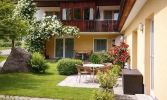 A cozy outdoor area with a table and chairs in a garden. In the background, there is a sunny building with flowers and trees.