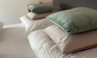 A cozy bedroom with a bed decorated with pillows in soft colors. In the background, there is a red wall design with a floor lamp visible.
