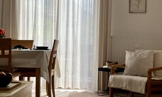 A bright living space with a dining table, chairs, and large windows. The curtains let in soft light and create a cozy atmosphere.