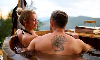 A couple relaxes in a wooden bathtub with a view of the mountains. The sunset creates a romantic atmosphere.