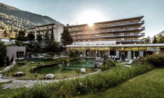 A modern hotel building with large windows and a spacious green area. In the foreground, a harmonious garden with a pond can be seen, surrounded by trees and mountains.