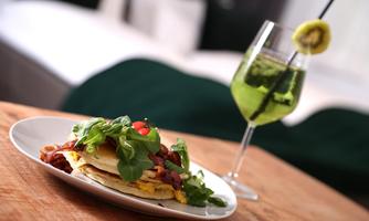 A plate with a hearty dish that includes pancakes, bacon, and fresh vegetables. Next to the plate is a green cocktail in an elegant glass.