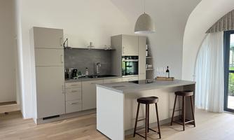 A modern kitchen with bright furniture and a large window. The room is designed with wooden flooring and minimalist elements.