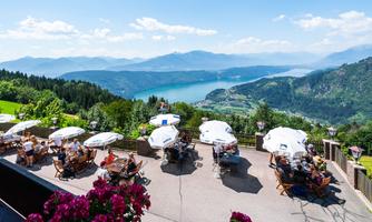 A sunny terrace with guests enjoying the view. In the background, a clear lake stretches out along with the surrounding mountains.