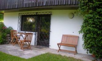 A cozy outdoor area with a table and chairs made of wood. The wall of the house is white, surrounded by green grass and plants.