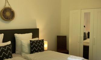 A bright bedroom with two single beds and modern pillows. In the background, there is a wardrobe and a light source.