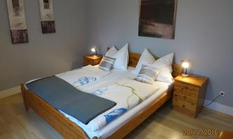 A cozy bedroom with a large double bed and light wooden furniture. Some pictures hang on the walls, and there are nightstands with lamps.