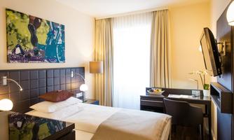 A modern hotel room with a single bed and a desk. The walls are light, and a large window lets in plenty of light.