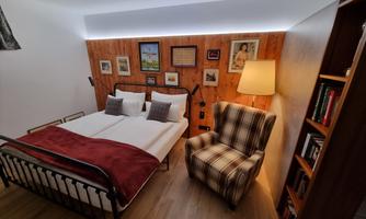 A cozy bedroom with a double bed and an armchair. Pictures hang on the walls, and there is warm lighting.