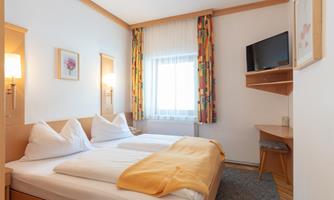 A bright and cozy bedroom with two beds. The room includes a desk, a television, and colorful curtains.