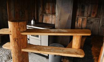 A rustic room interior with wooden shelves and a small stove. The walls are made of wood, creating a warm atmosphere.