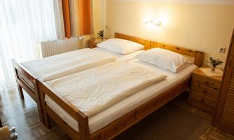 A cozy bedroom with a wooden double bed, white bedspreads, and a bedside table. Bright curtains let in plenty of light.